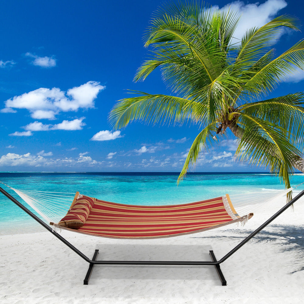 Red outdoor hammock with detachable pillow, set up on a beach for relaxation under palm trees and blue skies.