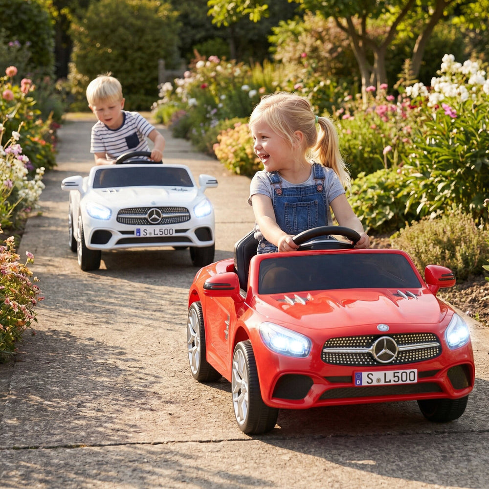 Two kids driving licensed Mercedes-Benz ride-on cars in a garden, featuring red and white models with LED headlights.