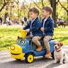Two children ride on a blue 12V Chuggington train with detachable carriage in a park, accompanied by a small dog.
