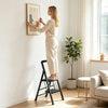 A person uses a 2 step black folding step stool with wide anti-slip pedals for stable climbing while hanging a picture.