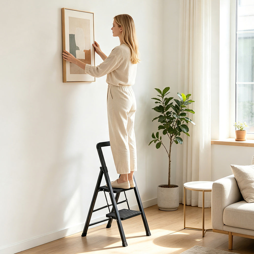 A person uses a 2 step black folding step stool with wide anti-slip pedals for stable climbing while hanging a picture.