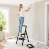 A person using a 2 step black folding step stool with wide anti-slip pedals to paint a wall in a bright room.