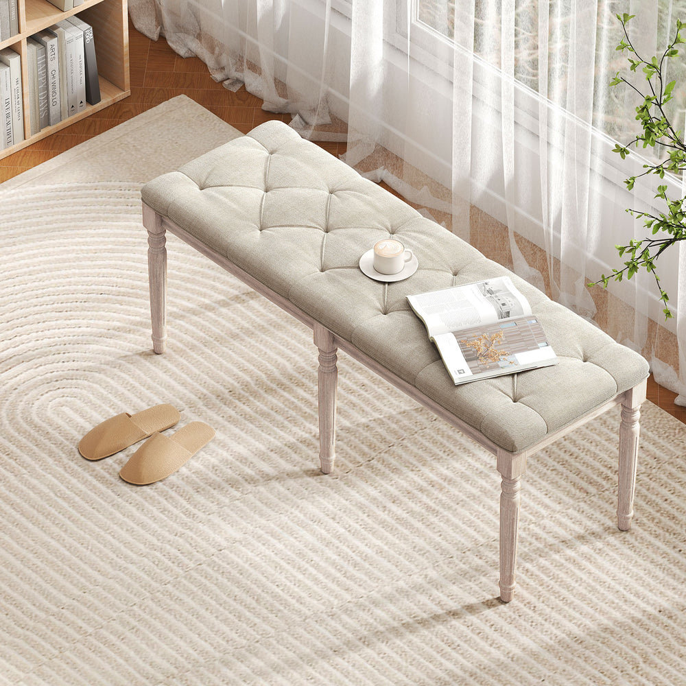 Beige upholstered bench with carved rubber wood legs, button-tufted seat, ideal for entryway or living room seating.