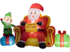 Christmas inflatable elf beside Santa Claus on sofa with colorful gift boxes, ideal for charming holiday displays.