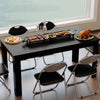 Black dining table with griddle grill, surrounded by folding chairs, perfect for six-person meals in compact spaces.