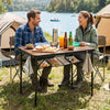 Folding camping table in black and brown, set up by a lake with drinks and snacks, featuring adjustable heights and.