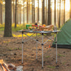 Folding roll-up camping table with metal legs, set up outdoors with food and drinks, perfect for picnics and camping.