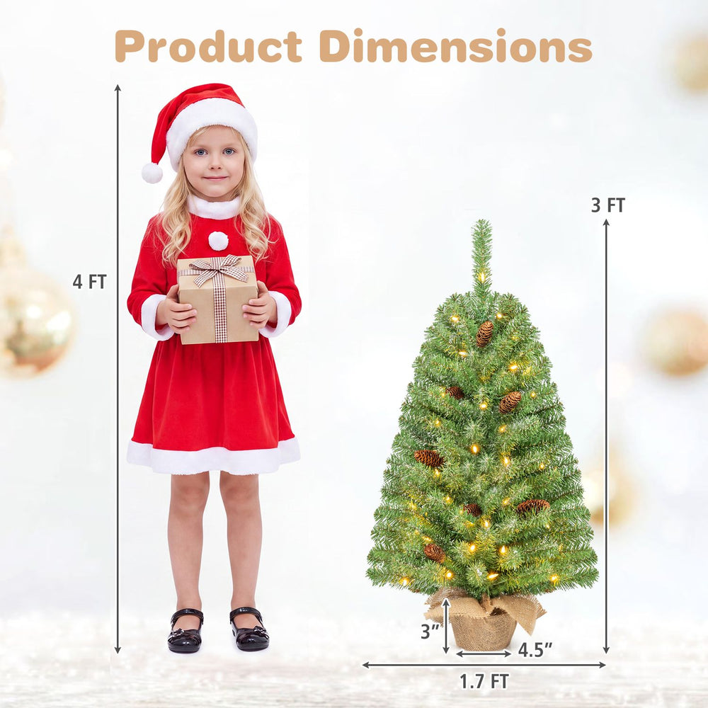 Pre-lit mini Christmas tree with 50 warm white LED lights, pine cones, and a linen-wrapped base, shown beside a child for scale.
