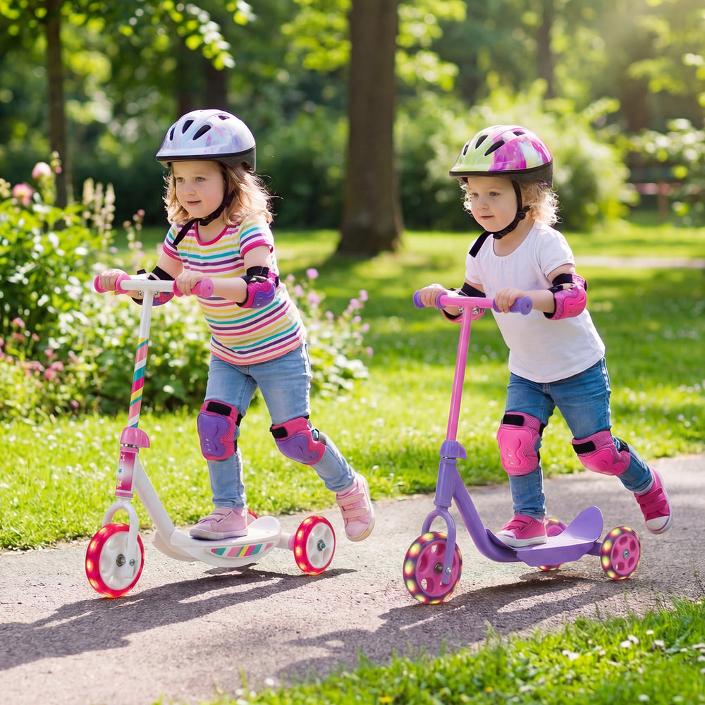Two toddlers riding pink kick scooters with LED light-up wheels on a path in a sunny park, promoting balance and fun.