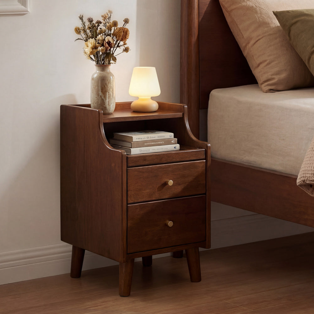 Wood nightstand with two drawers and solid wood legs in brown, styled beside a bed with decorative lamp and books.