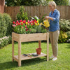 Wooden raised garden bed with legs, filled with colorful flowers, featuring a storage shelf for gardening tools.