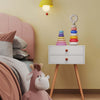 White storage end table with two drawers beside a pink twin bed and colorful stacking toy in a children's room.